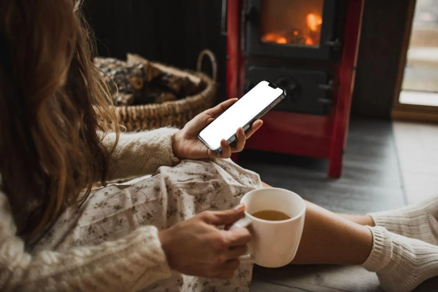  Womsan sat looking at a phone. She holds a cup of tea and is sat on the floor in front of a log burner.