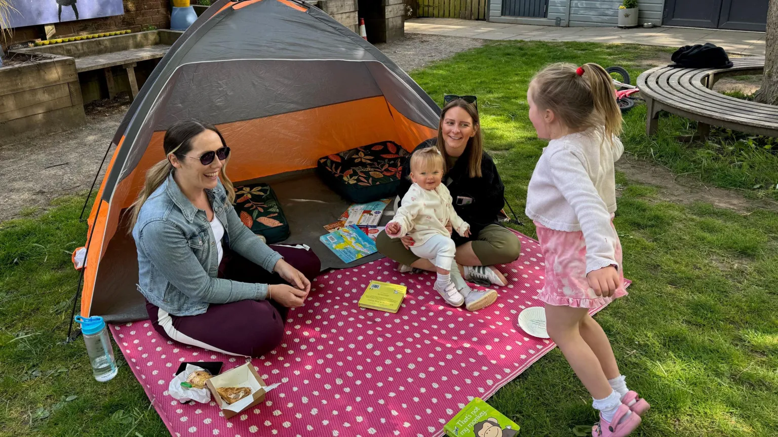 Include Us Two mums with children sit outside a tent. One of the children is sitting on her mother's knee, the other is jumping in the air.