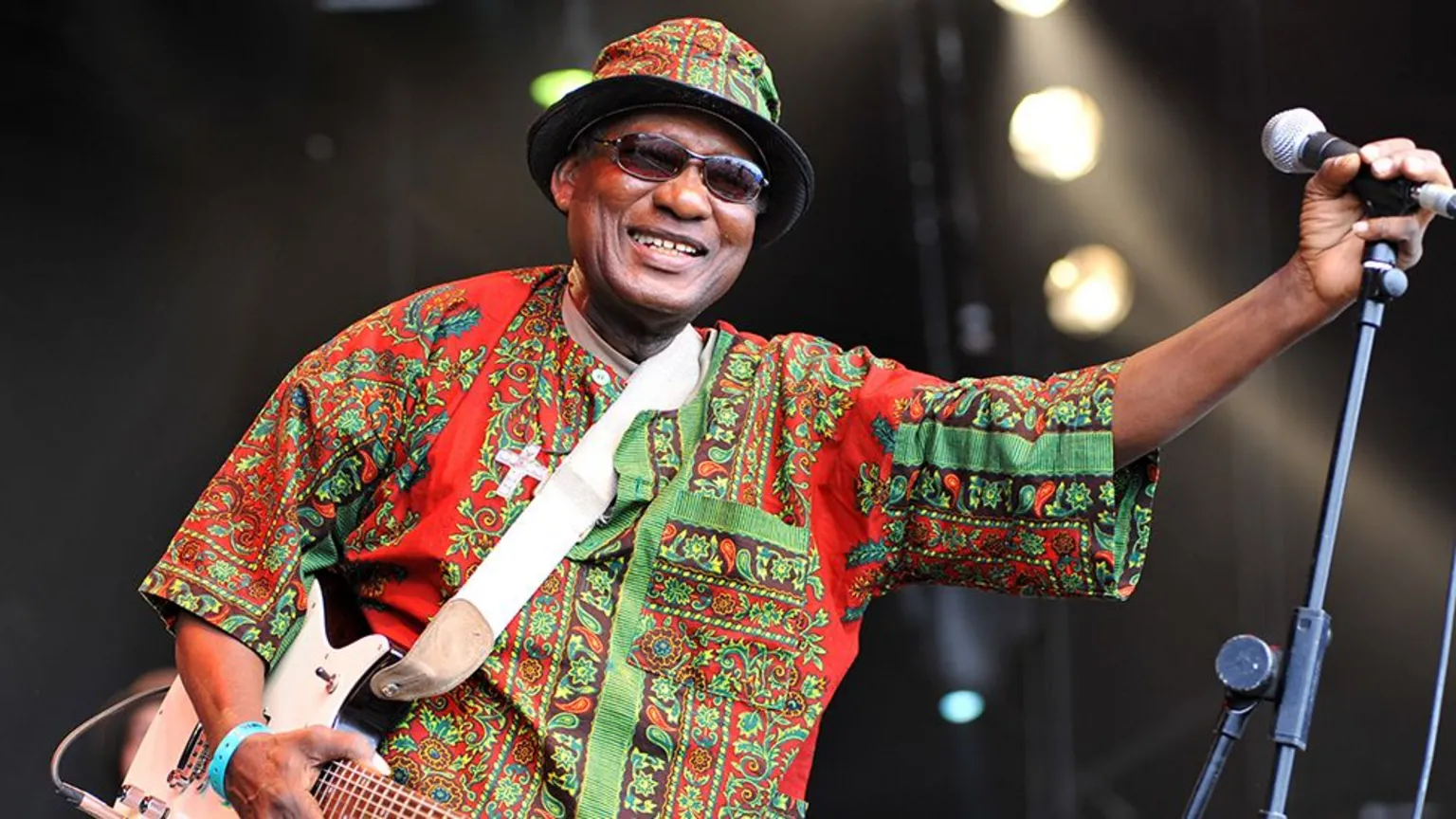  Ebo Taylor in a colourful African shirt and hat and dark glasses. He is smiling and holding a microphone 