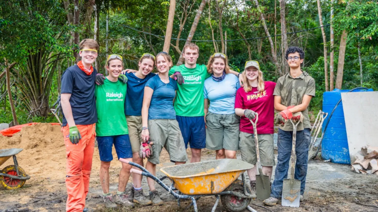 Katie Henderson/Raleigh International A group of young people standing in the jungle with their arms round each other. There appears to be a mound of sand behind them and there is a yellow wheelbarrow in front of them. Some have spades and goggles and they are wearing shorts, t-shirts and boots or trainers. Their legs are spattered with mud and they are smiling. 