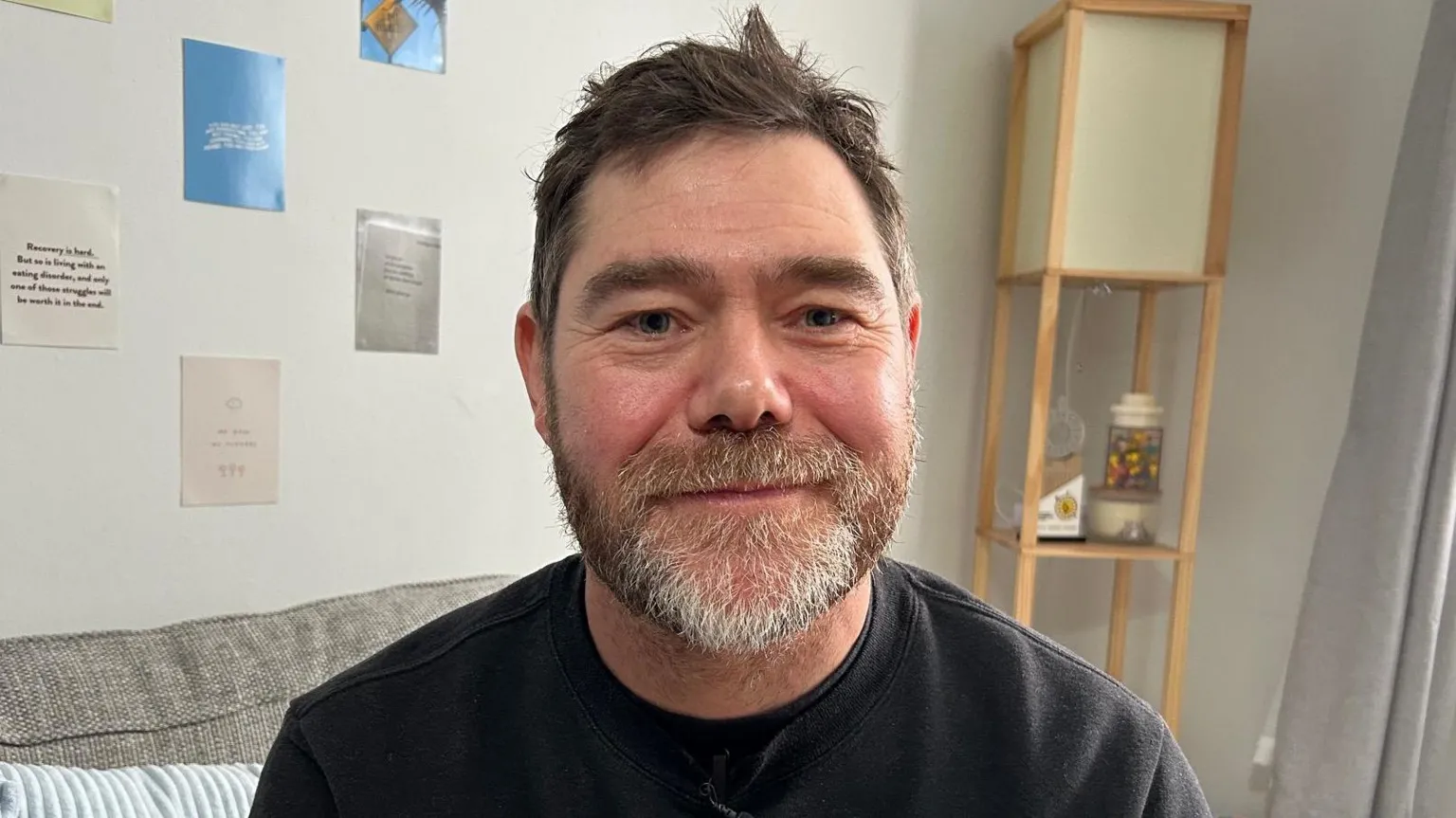 Stephen has dark brown hair and a greying goatee beard. He is wearing a black jumper and is smiling at the camera. He is pictured on a sofa in Milly's home. 