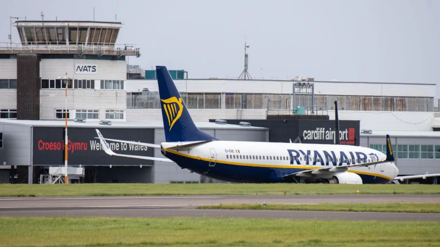 A Ryanair plane, with white body, blue text and a blue and gold harp logo on its tail fin, taxis in front of the terminal building at Cardiff Airport. Grass and tarmac can be seen in the foreground.