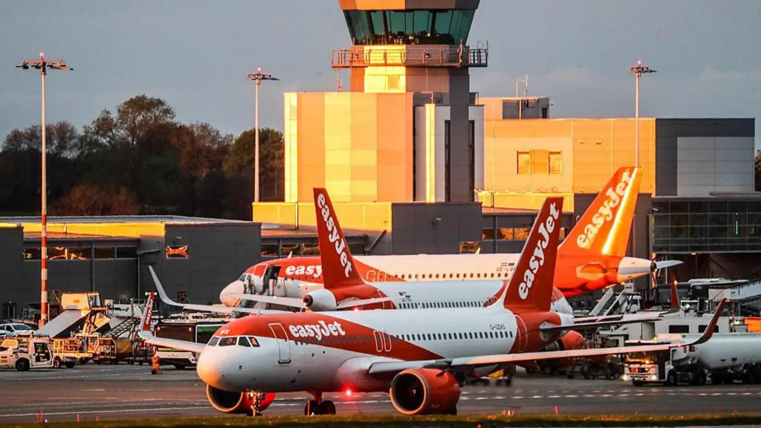 Three planes with EasyJet orange and white branding are on the tarmac at Bristol Airport, with the terminal building in the background bathed in the golden light of the setting sun.