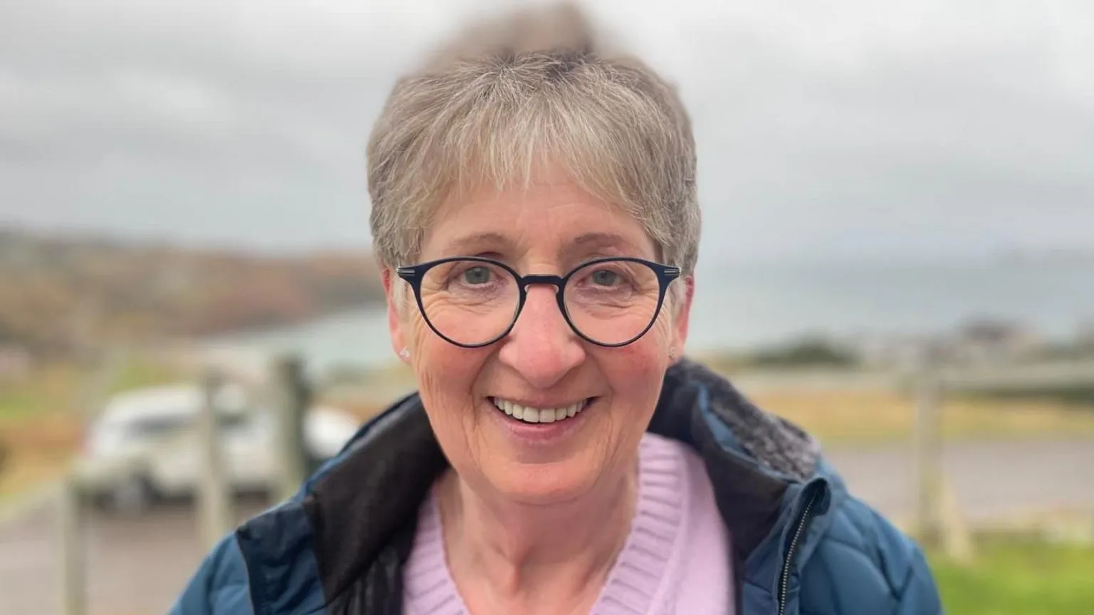 Margaret Mackay looking directly at the camera. She has short hair and black rimmed glasses and is wearing a pink jumper and a teal jacket. In the background is a fence, a white car and a coastline.