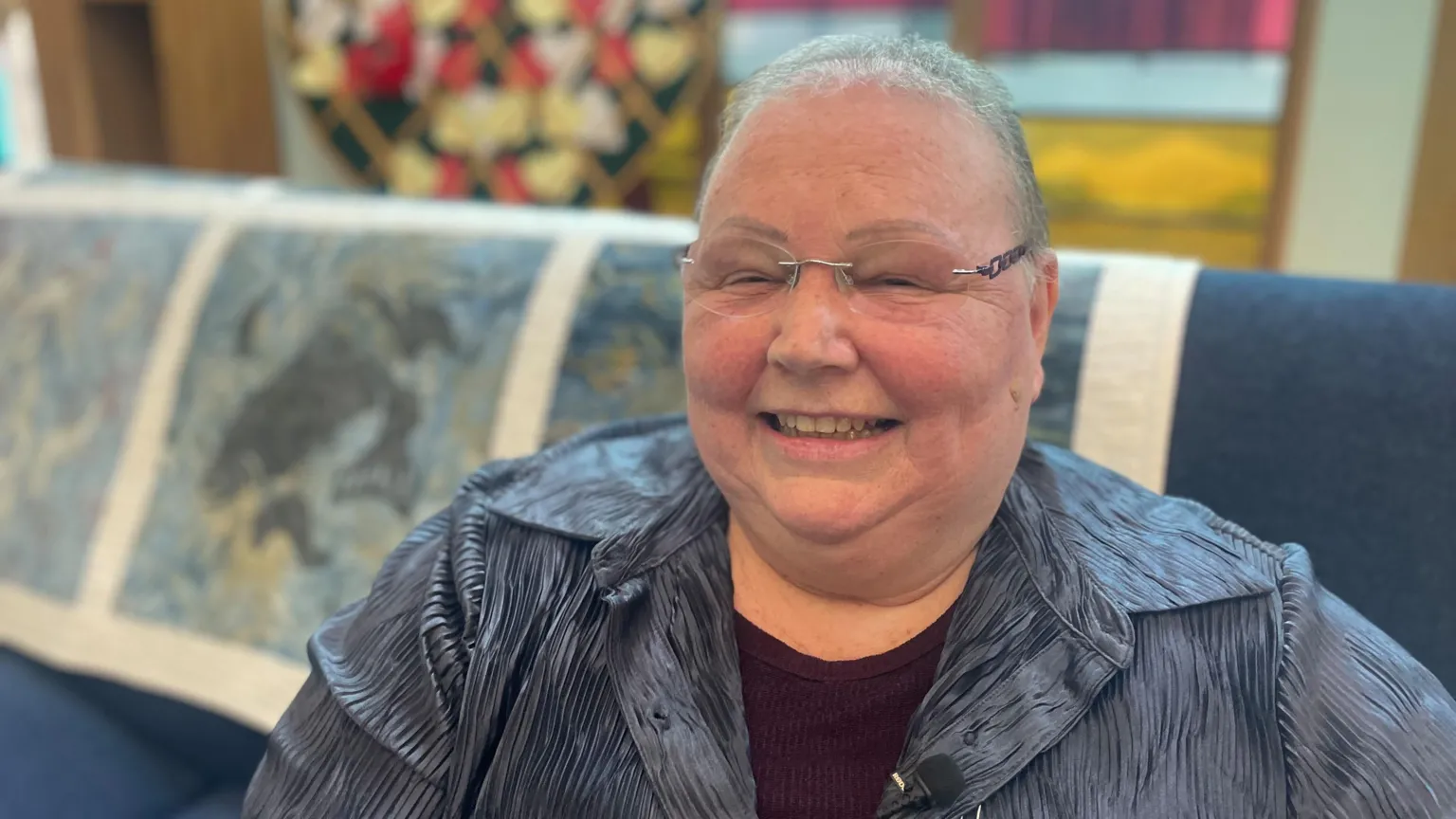 Karen sitting on a blue couch with a patterned throw over the back. She has short hair and glasses and is wearing a blue blouse and a burgundy top.