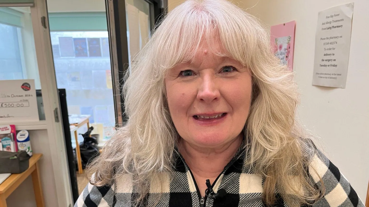 Andrea looking directky at the camera in a rom with white walls and an open door behind her. She has long white hair and is wearing a black and white checked top with a zip.
