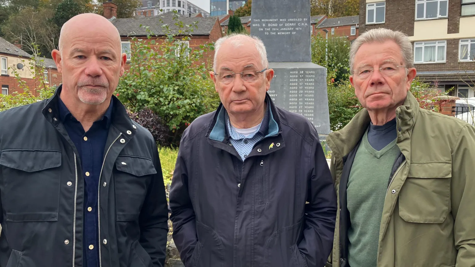 Three men stood looking at the camera. On the left is a bald man wearing a navy-blue shirt and a black jacket. In the middle is a man with glasses wearing a dark jacket. On the right is a man with grey hair wearing a green jumper and a green jacket. The appear to be standing in front of a monument.