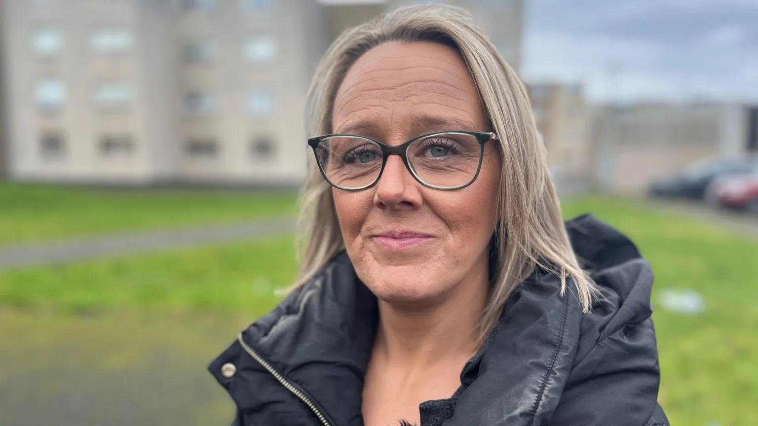 A woman with blonde hair and dark rimmed glasses in front of a block of flats.
