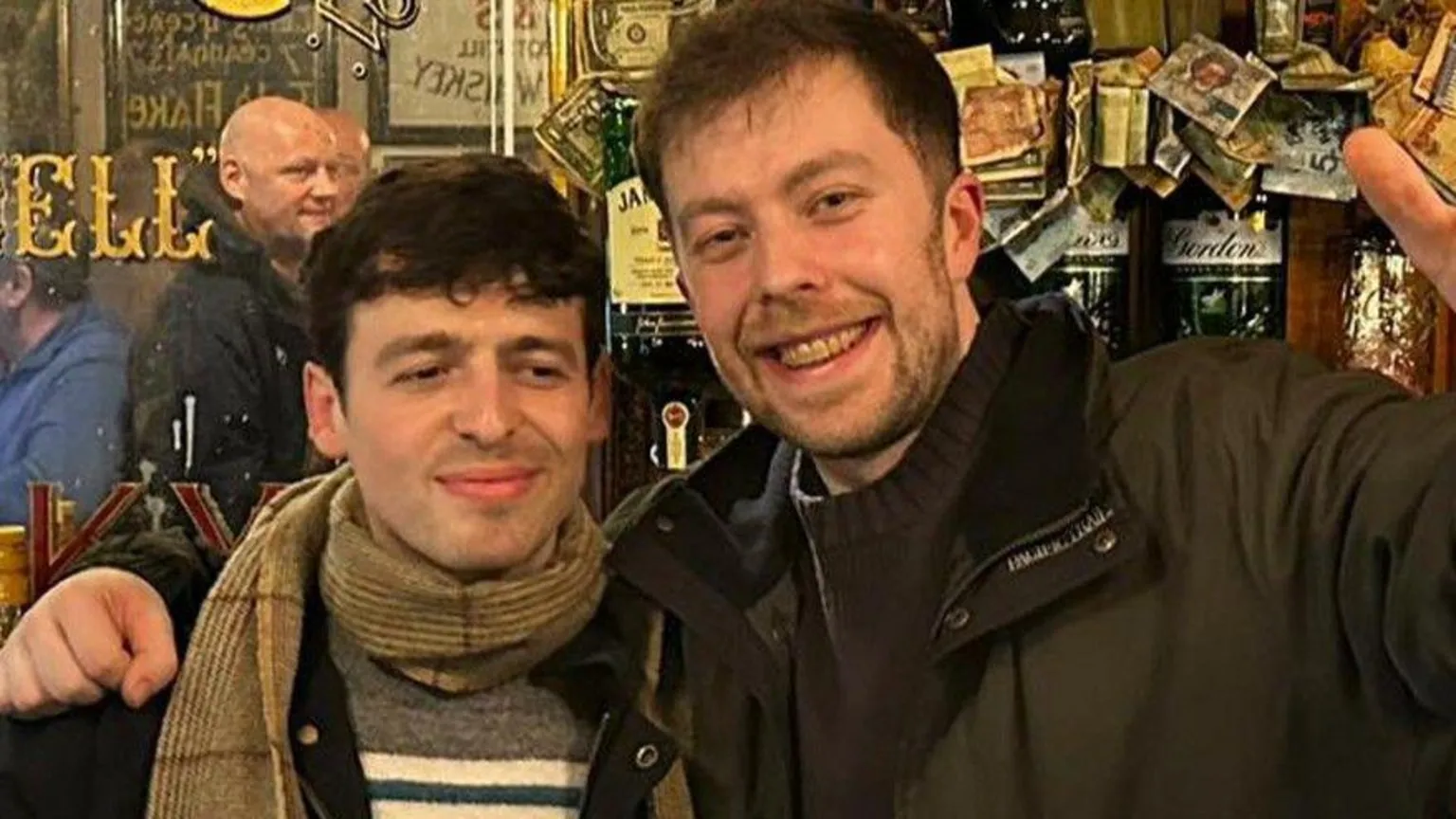 Peadar O'Donnell's Two men stand in a pub in front of a long mirror and the bar. They are wearing winter coats. The man on the right has one arm held aloft.