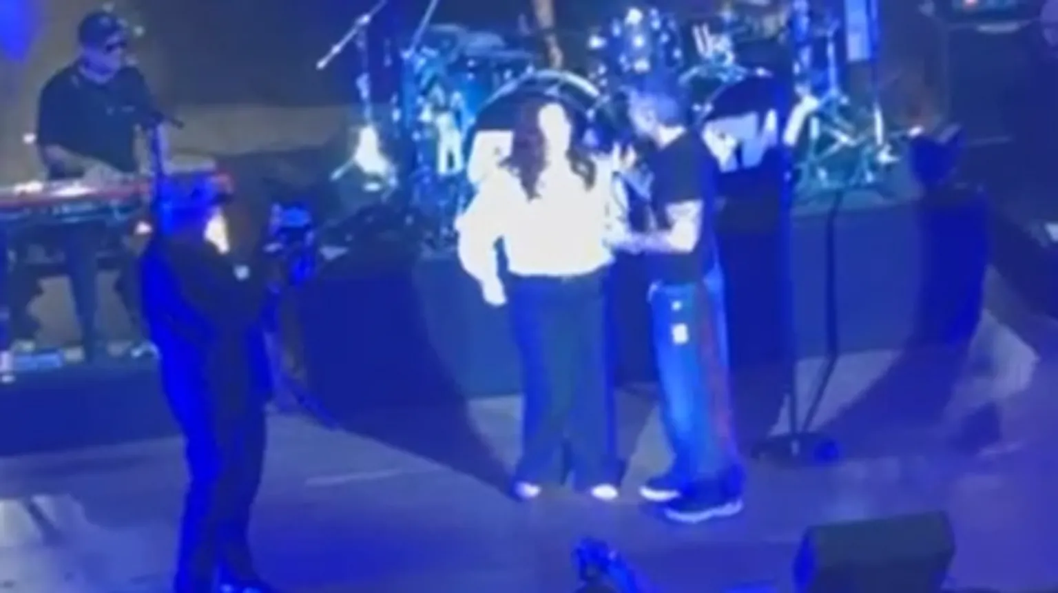 A zoomed in image of a woman in a white top and dark trousers standing next to a Robbie Williams, a man in a black tshirt and jeans on stage. Behind them there is a set of drums and on the left of the image is a man in a black tshirt, cap and sunglasses playing the keyboard. In front of him is a man in black holding a camera