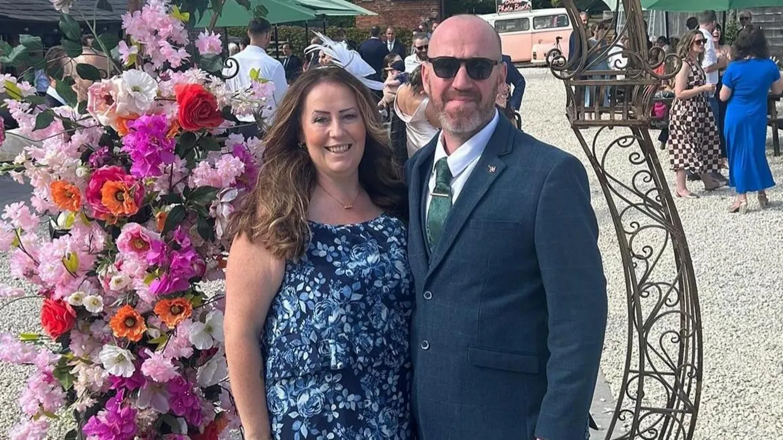 Jemma Williamson Jemma Williamson, a woman with wavy brown hair in a blue flowery dress is smiling at the camera and standing next to a man in a blue suit with a green tie and sunglasses. Behind them in a flowery display 