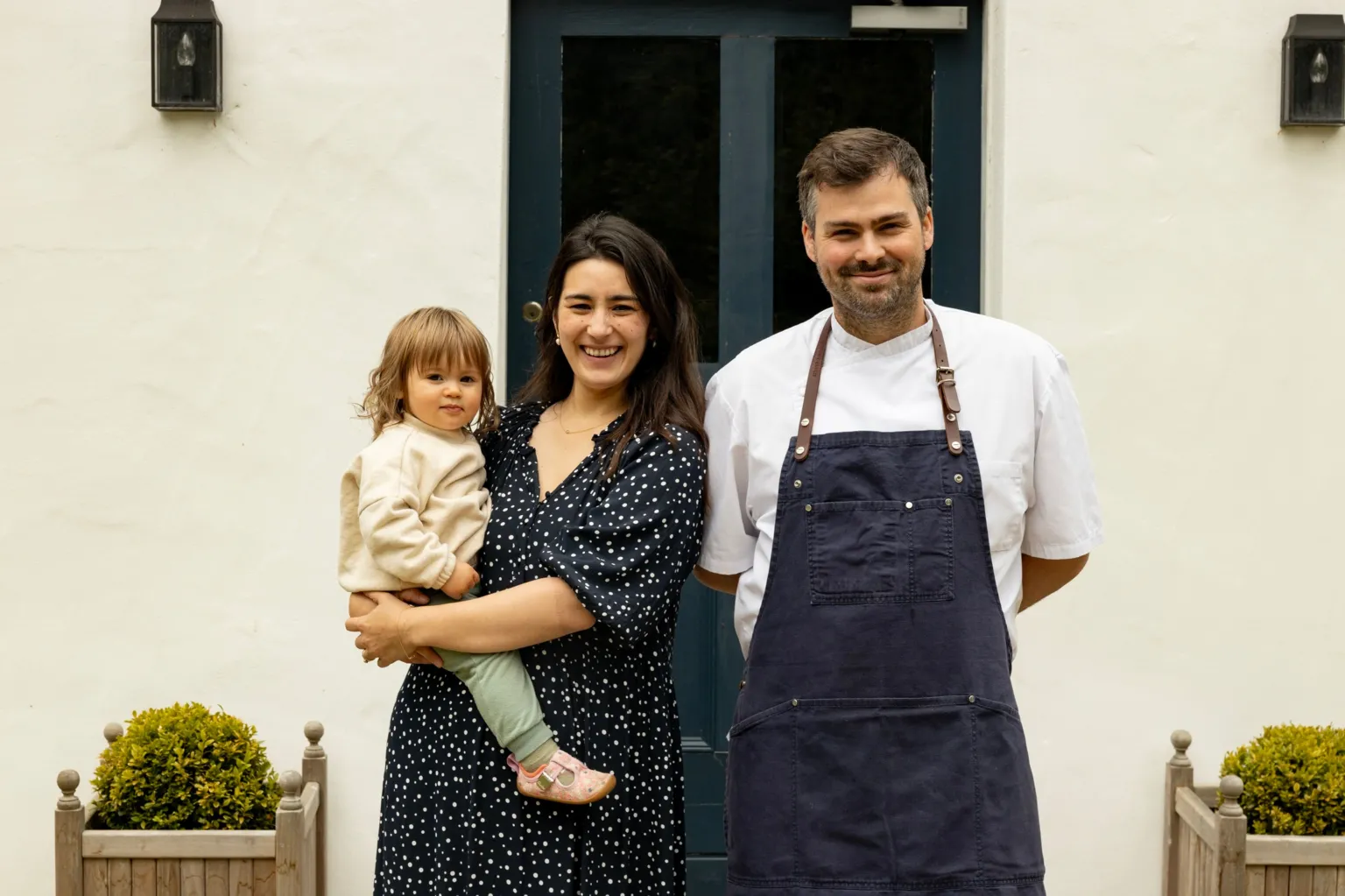 Alex Baxter Photography Matilda and Tom Tsappis standing outside Killiecrankie House. Matilda has shoulder length dark brown hair and is wearing a black dress with white spots and is holding a toddler on her hip. Tom has short brown hair and a trimmed beard. He is wearing a white chef's jacket and a navy blue apron with leather neck strap.