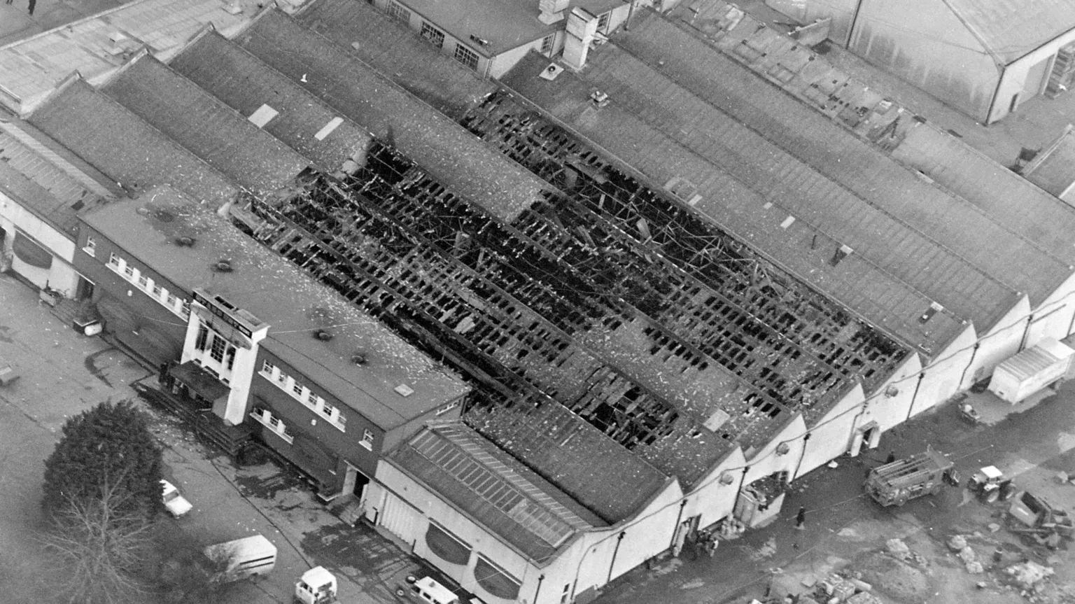 Independent News and Media/ A black and white aerial shot of Stardust nightclub after the fatal fire. The large, warehouse-like complex consists of several attached buildings with pitched roofs and a main entrance with a flat roof and large windows. More than a third of the pitched roofs are badly burned, with damaged rafters visible.