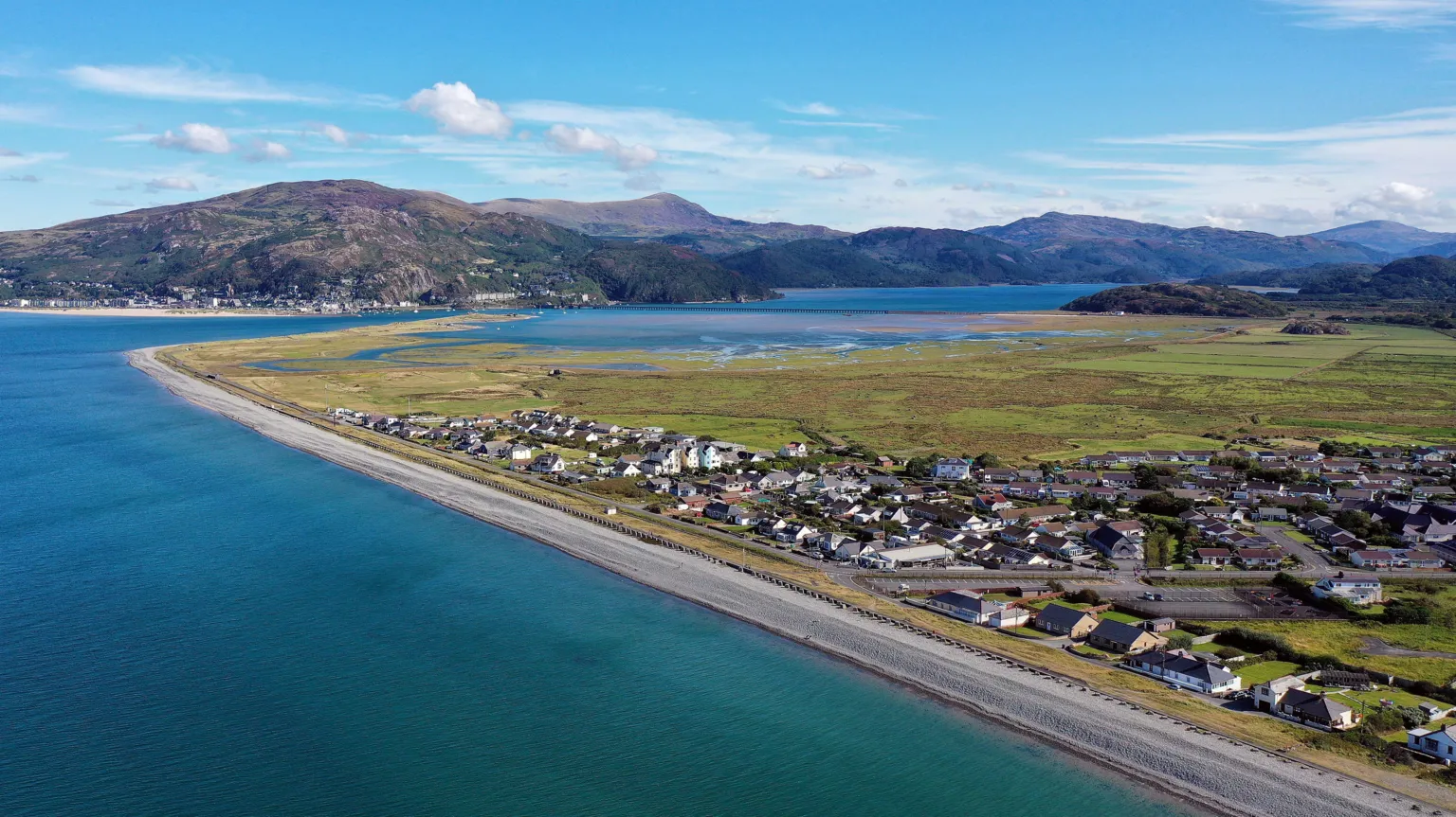  An ariel shot of Fairbourne. The village is on the coast, which curves around, and houses and roads can be seen with a stretch of grass behind it. A mountain range can be seen in the horizon. 