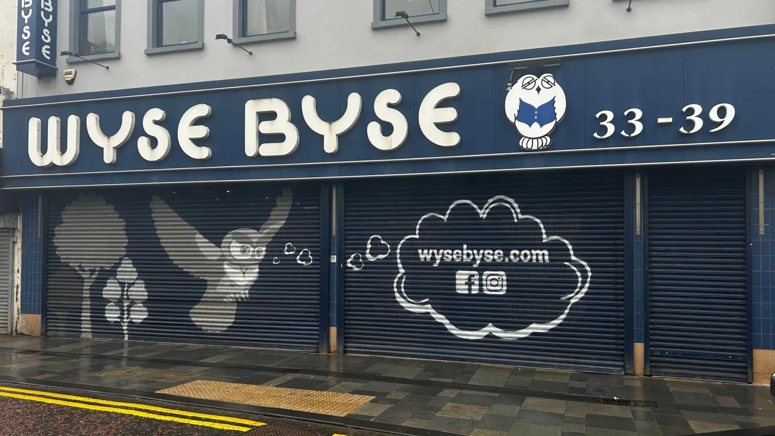 Wyse Byse store front with blue shutters down. The sign WYSE BYSE is in white and the shops mascot is an Owl painted beside it. The white owl wears a black graduation cap and holds a blue book. 