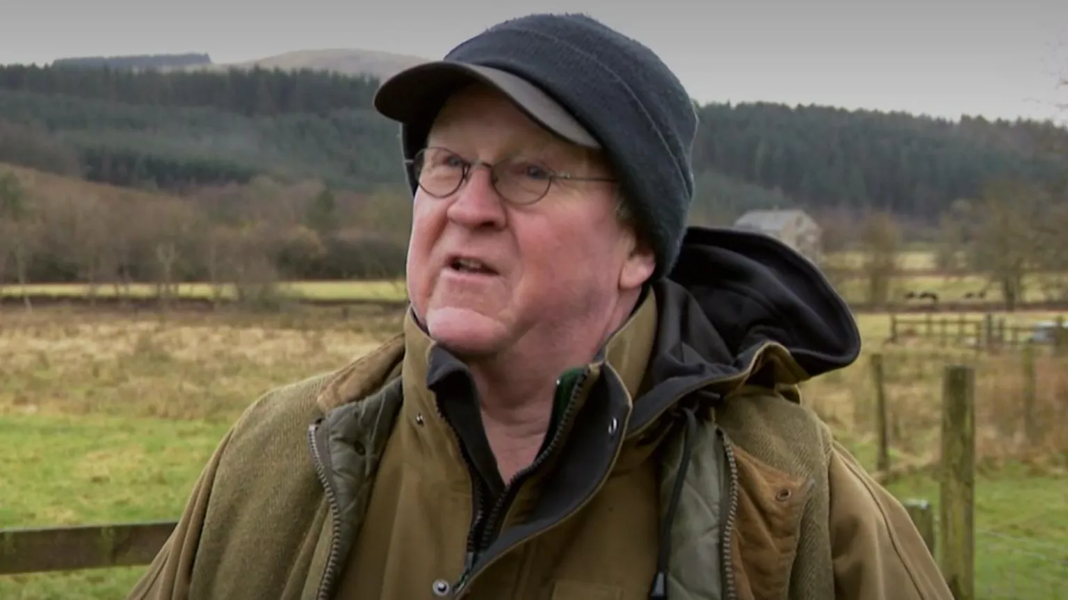 A man in a black hat with glasses and a green jacket stands in front of a fence with tree-lined hills in the background