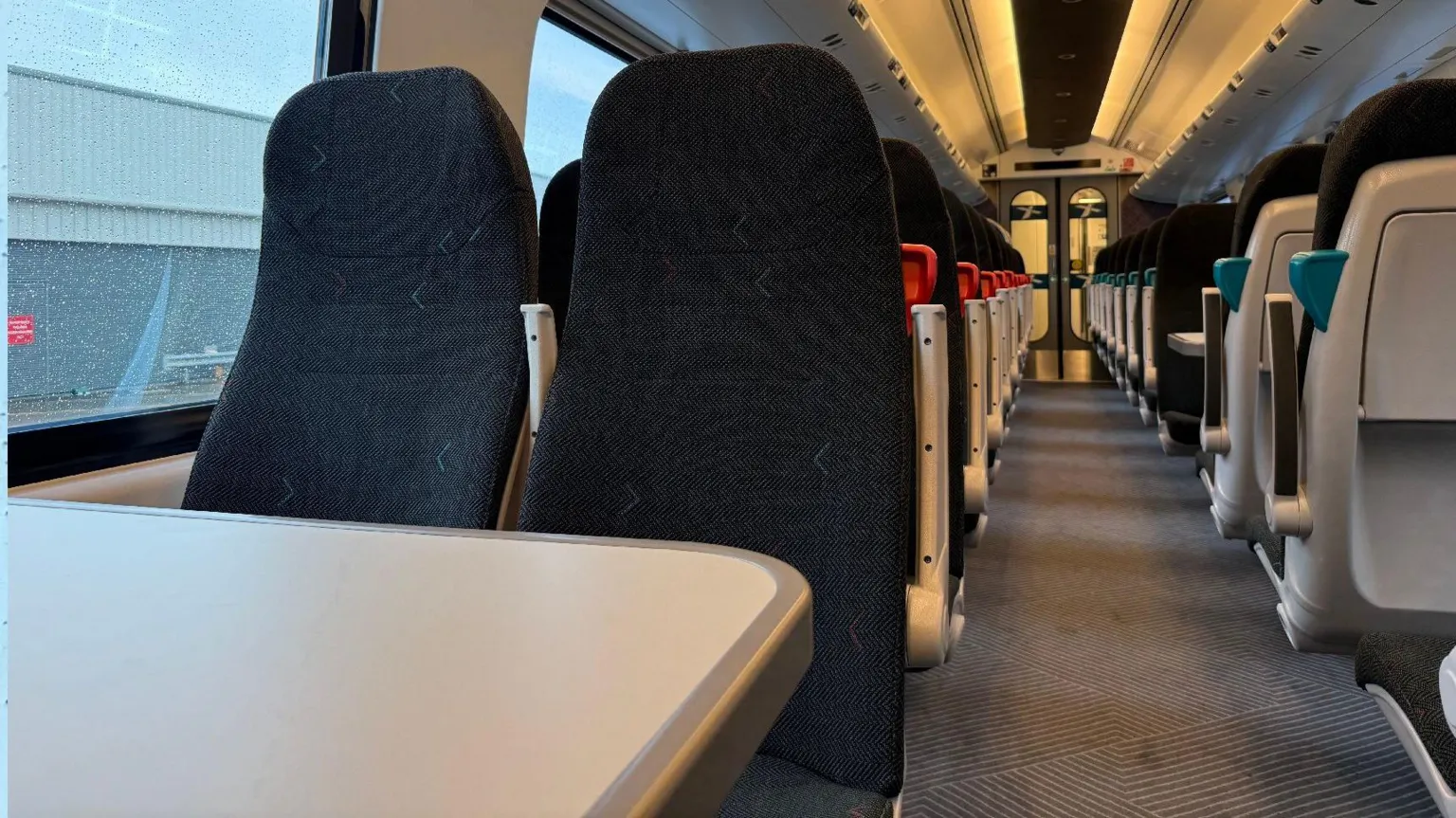 CrossCountry Image shows the inside of a refurbished CrossCountry train carriage. The carriage is empty and has black fabric seats with grey tables,
