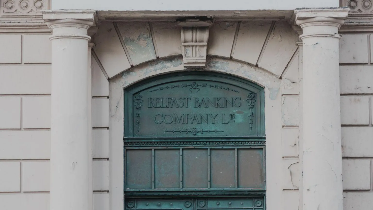 The World Monuments Fund A green metal sign says Belfast Banking Company. Two columns are on each side of the door. 