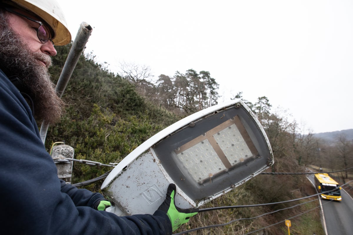 A man in a hard hat holds the top of a streetlight. Far below a bus can be seen driving along the road