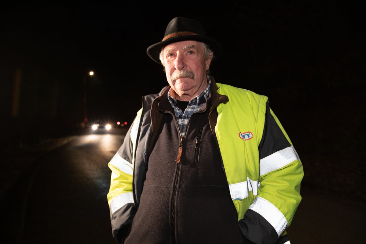 A man in hi-vis and a hat stands next to a road at night