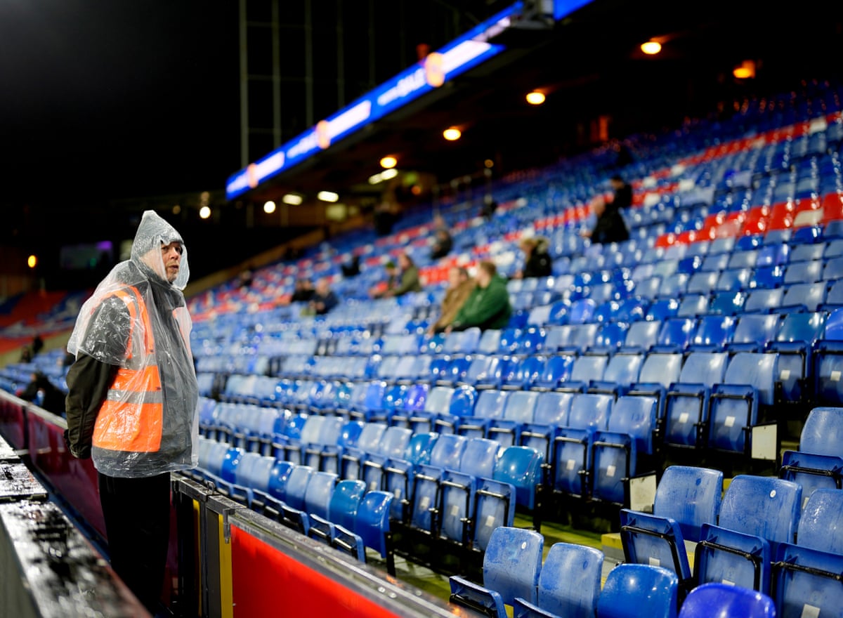 A steward wearing a waterproof poncho in front of a smattering of fans ahead of the Premier League match between Crystal Palace and Burnley at Selhurst Park.