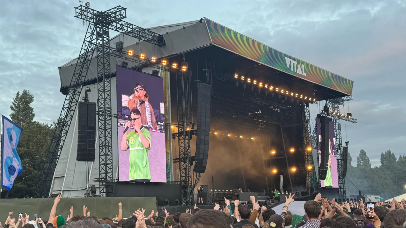 A large pop up stage outside. It has two speakers on either side and two large screens displaying images of Irish rap trio Kneecap who are performing. A large number of people are gathered for the gig and many of them are holding their hands in the air.