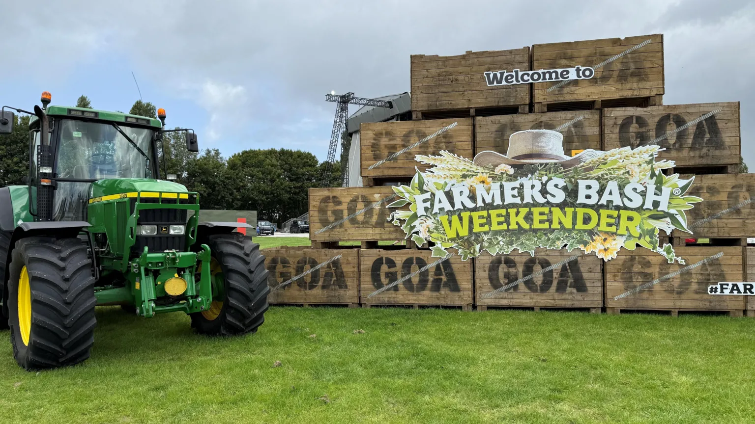 A large green tractor and brown crates sitting on green grass. The crates have advertising on their front. Trees and the corner of a stage are in the background. 