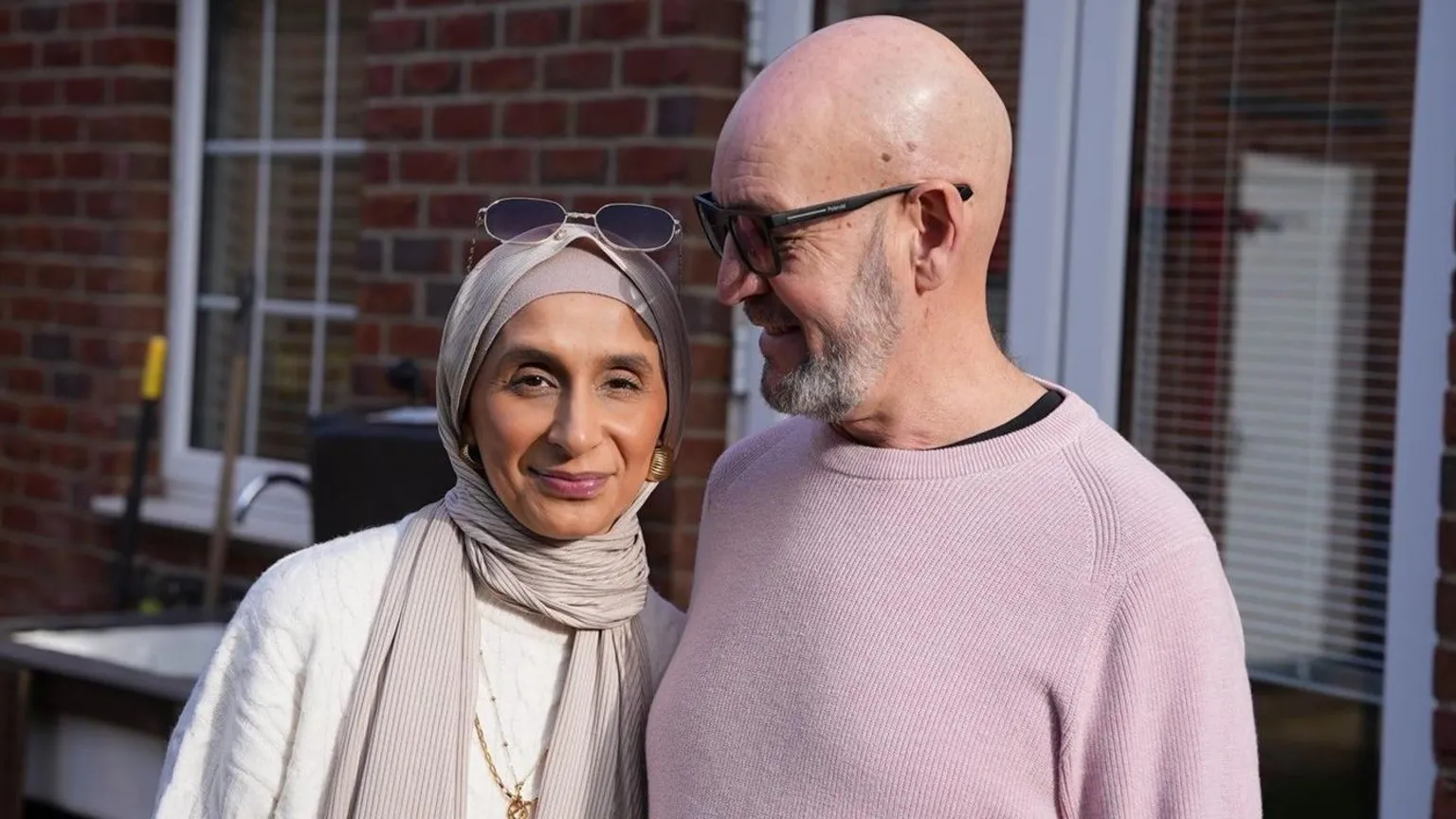 This photograph is of Sahir and Dennis in their garden. Sahir, on the left, is wearing a creamy jumper with a soft cream headscarf. She has a pair of glasses on her head and is wearing gold necklaces. Dennis has his arm around Sahir and is looking at her as she smiles at the camera. He's wearing a light pink/lilac knit jumper with a black t-shirt underneath. He's wearing glasses and has a beard. 