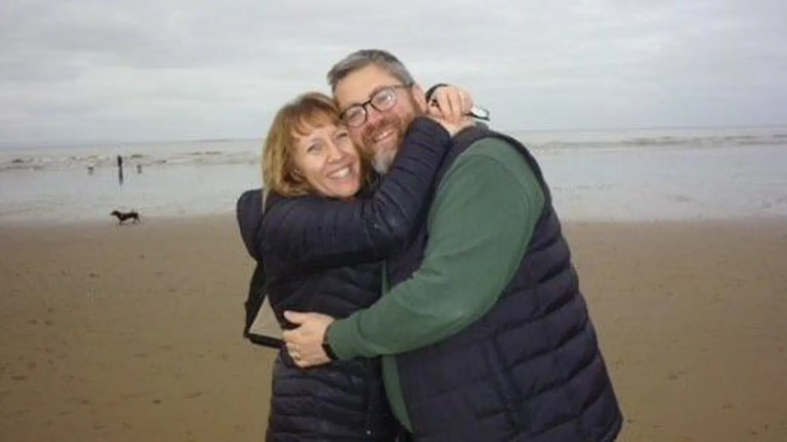 Carl Hickling Carolyn, wearing a black puffer coat, hugs her husband - who wears a navy puffer and green jumper - on a windswept beach
