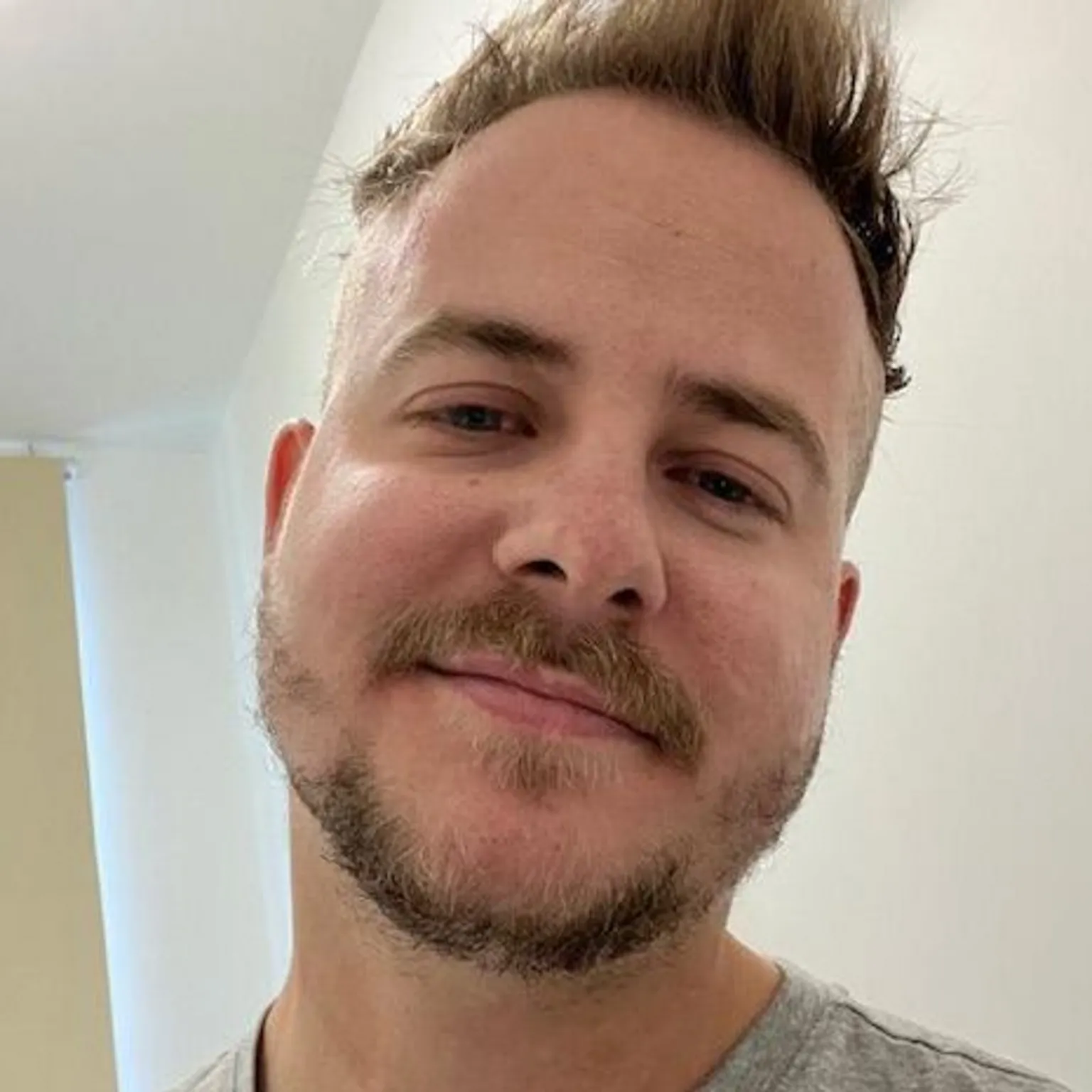 James Deegan James wearing a light grey T‑shirt standing indoors in a bright room. Part of a doorway or wall panel is visible behind him, with a window and a ceiling light in the background.