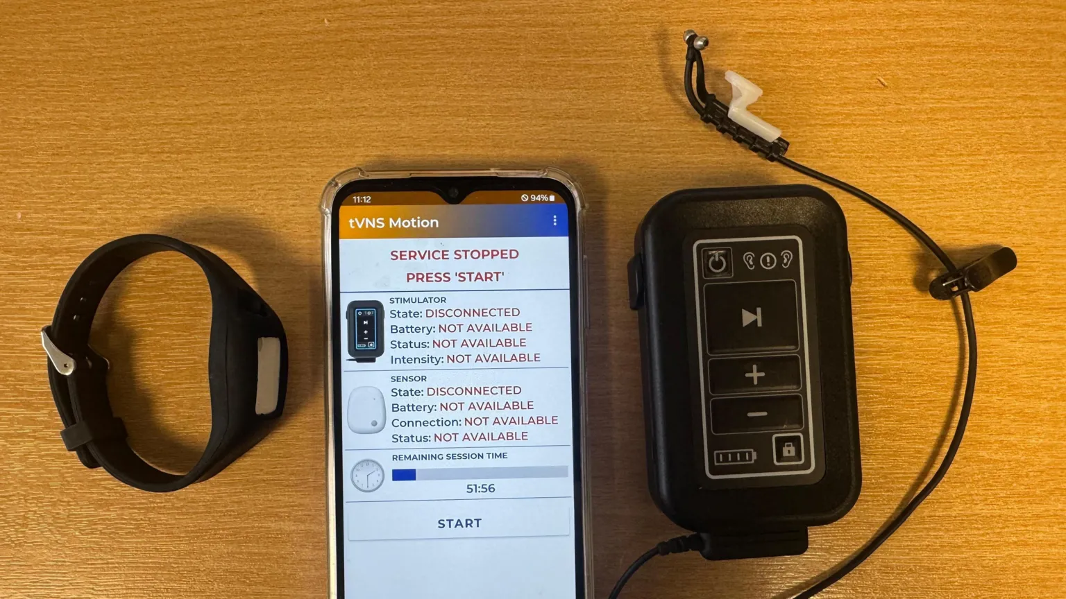 A watch, smartphone and the portable device with an ear piece on a table. The smartphone displays an interface to control the device, saying 