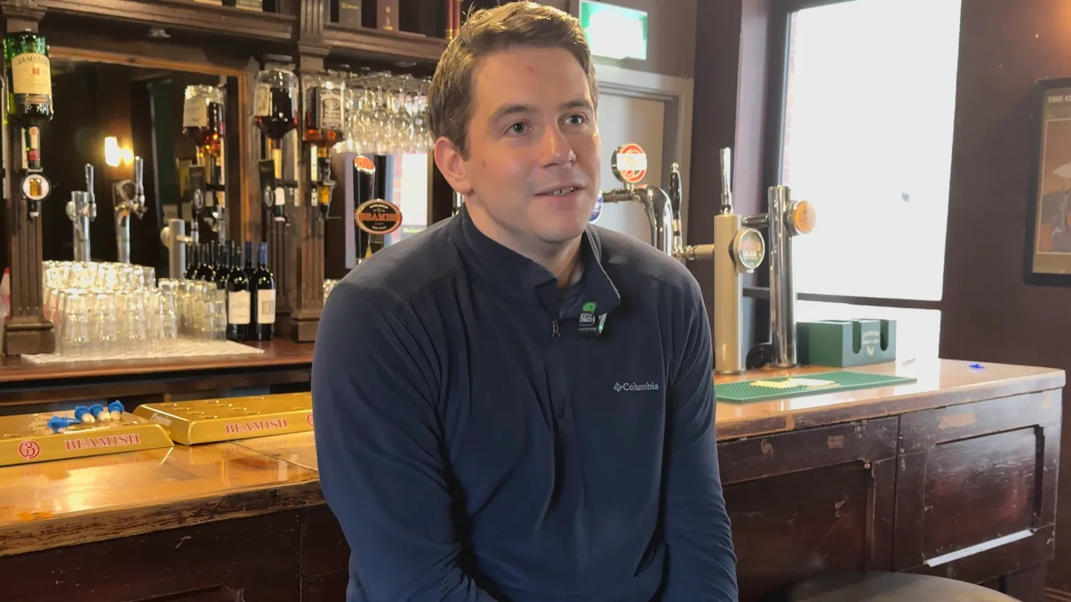 Cathaóir sits on a stool in front of a bar facing the interviewer. He has short brown hair, and is wearing a navy Columbia brand fleece. A small microphone is clipped to his fleece zip. Behind him is a bar, with taps featuring different pints on draught, as well as spirits hanging on optics.