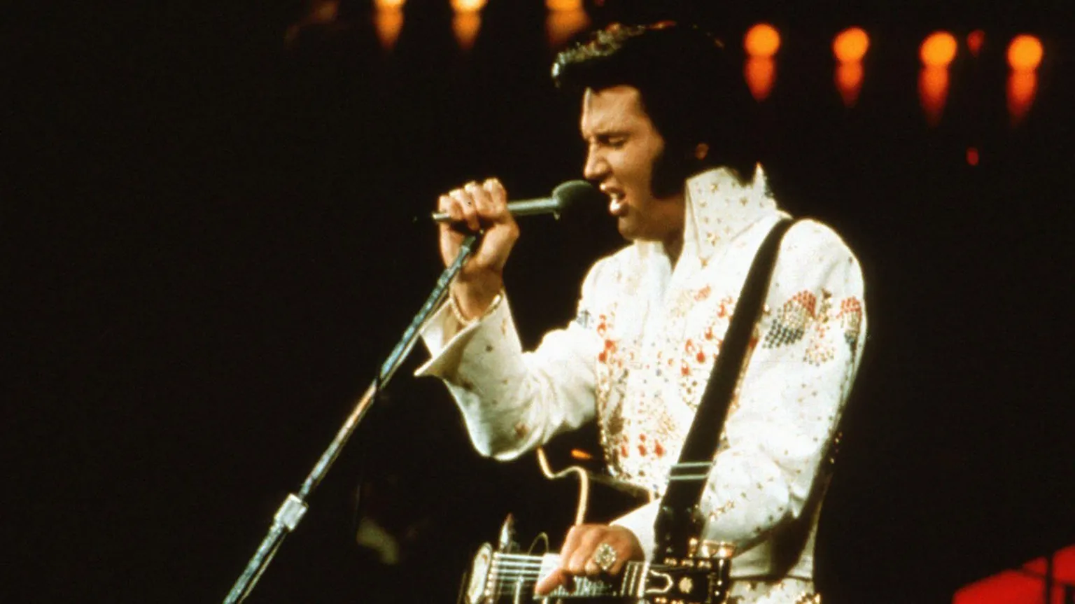  Elvis Presley is pictured on stage in 1974 wearing his iconic white outfit and dark hair spruced up. He is singing into a mic, with his acoustic guitar hanging around him by a strap.
