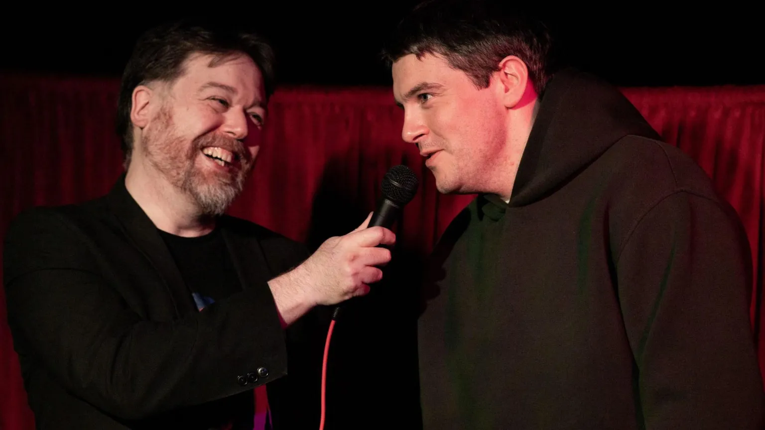 Brendan O'Connor Two men backed by a silk red curtain behind them. One on the left has a greying beard and short dark hair dressed in all black. He holds up a microphone with a red cable to the man on the right, who has short brown hair. He is wearing a dark green hoodie.