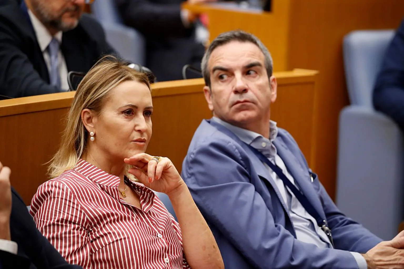  A woman in a red and white blouse with medium-length brown hair with streaks of blonde sits with her arm up towards her face in the Italian parliament. Next to her is a man in a blue suit with dark hair looking over his shoulder