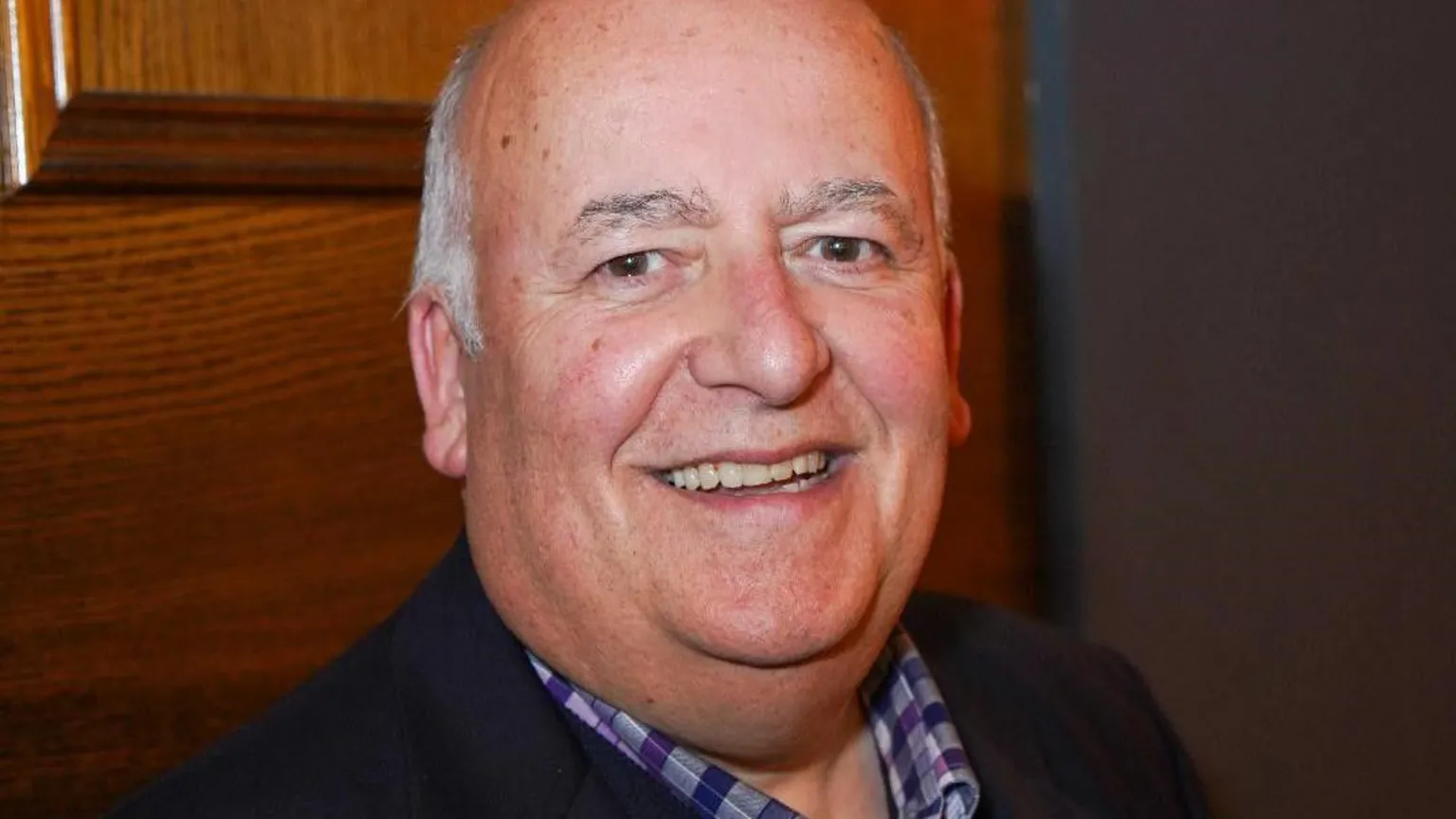 Raffaello Gonnella A bald headed man with short grey hair and brown eyes smiles into the camera. He has a checked purple shirt on and a dark jacket. He is in front of a wood panel.