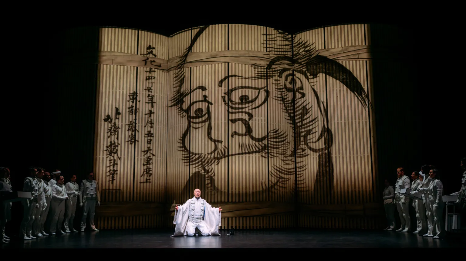Mihaela Bodlovic A man dressed in white kneels on the stage. A black ink drawing of a man with Japanese script is displayed in the background.