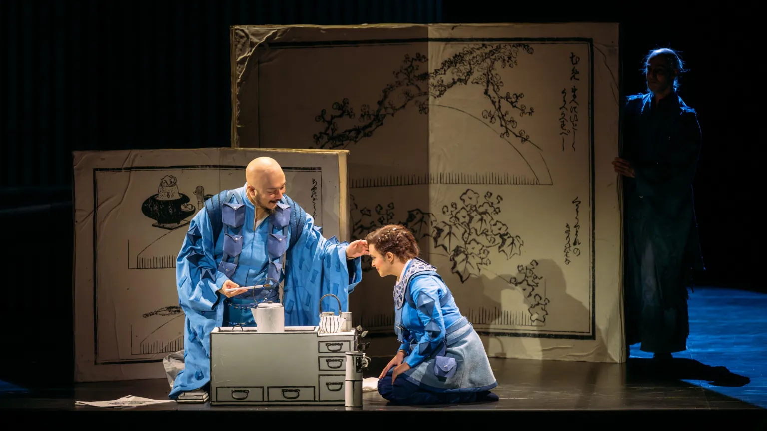Mihaela Bodlovic A man and a woman wearing blue robes kneel on the stage. There is a small kitchen unit on the stage and two boards in the background with black ink drawings, inspired by Japan.