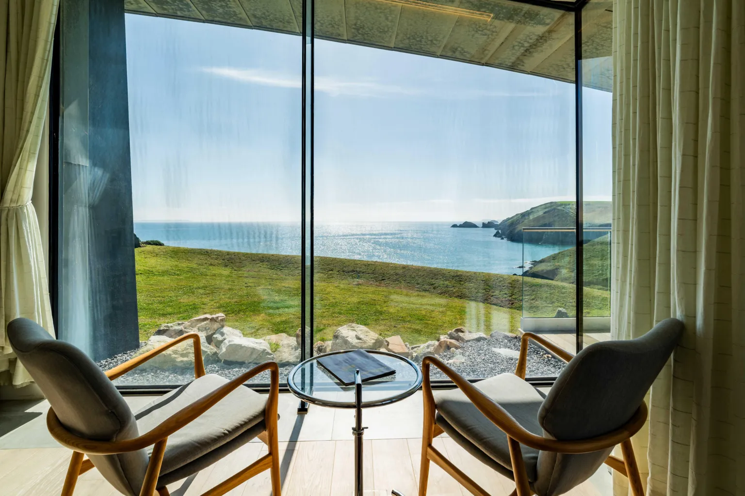 Fine & Country Two armchairs face a large floor-to-ceiling window, overlooking the sea with grassy cliffs in the distance.