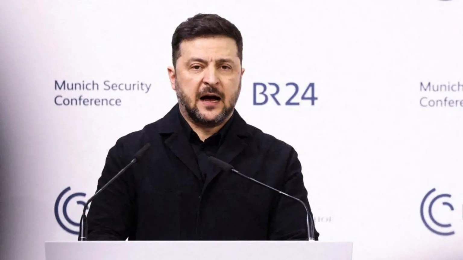  Volodymyr Zelenskiy in a dark coat stands at a podium with two microphones, speaking in front of a white backdrop displaying the Munich Security Conference and BR24 logos.