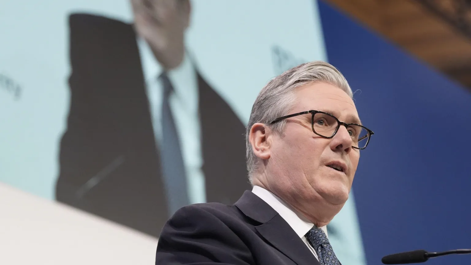  UK Prime Minister Keir Starmer talks into a microphone, wearing a dark navy suit jacket, dark navy and pink printed tie and a white shirt. His mouth is partially open and he is wearing a pair of dark-framed glasses. His image is projected onto the wall behind him. 
