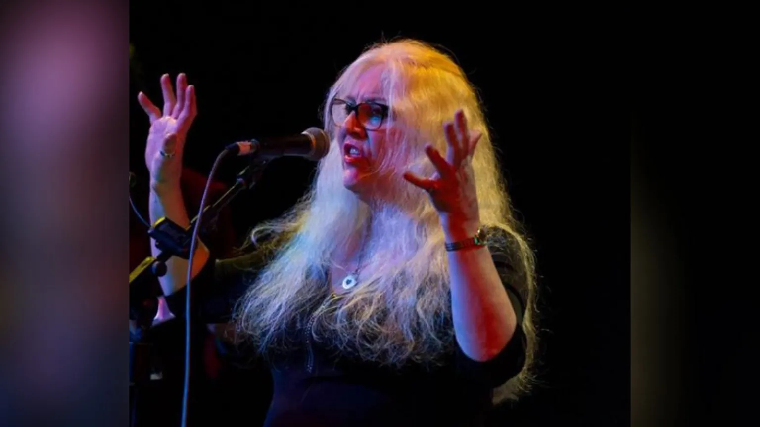 Marc Lynch Alison is onstage. She's singing in front of a mic with her hands up. She wears glasses and has long hair. She's wearing a silver necklace and a watch.