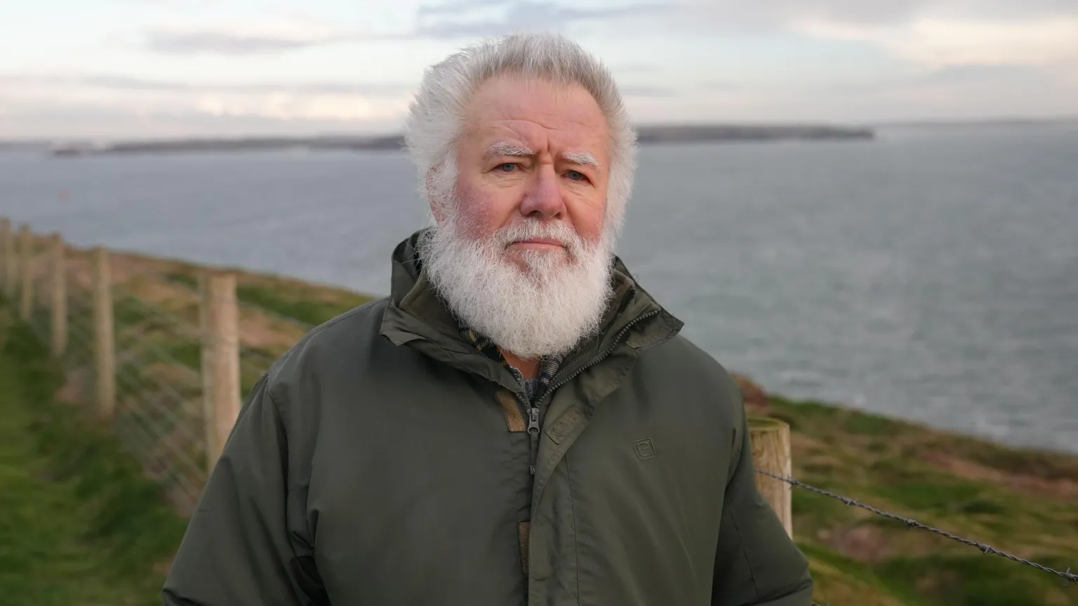 Retired BBC Wales reporter Hefin Wyn at St Anne's Head, Pembrokeshire. He has white hair and a white beard and is wearing a green, waterproof coat. Behind him can be seen the entrance to the Milford Haven waterway.