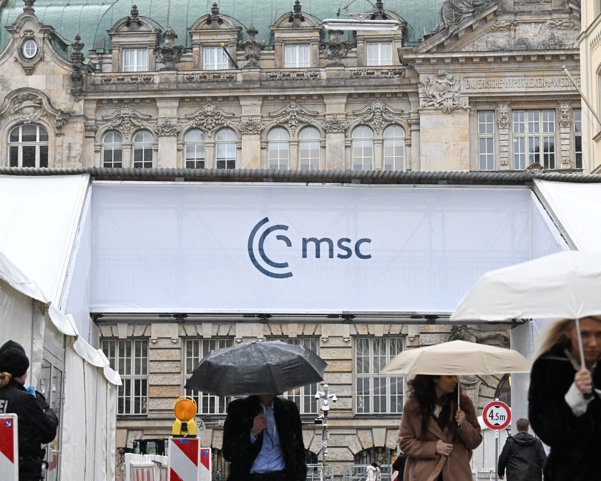 People with umbrellas walk past a pedestrian bridge with the logo of the Munich Security Conference leading to the hotel 