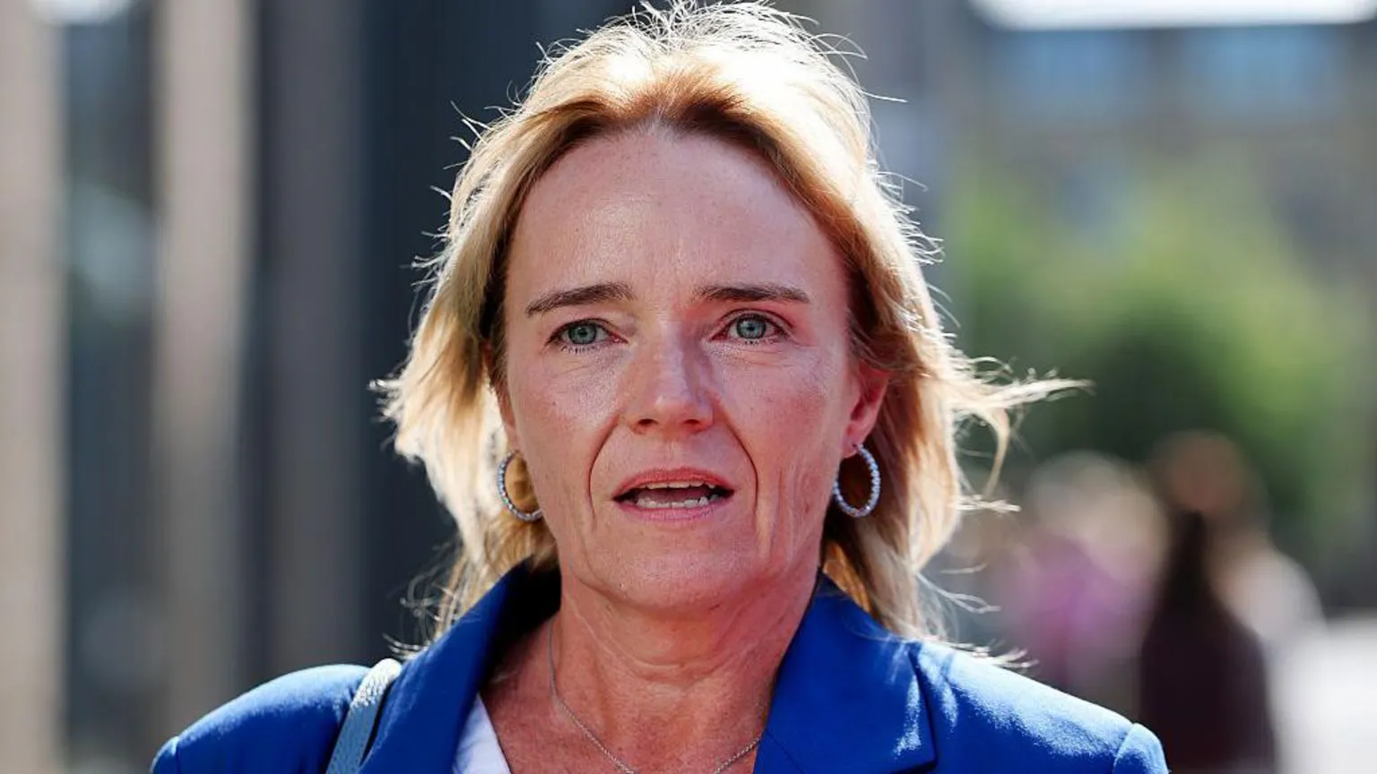 Sandy Peggie has shoulder-length blonde hair and is wearing a blue jacket over a white top