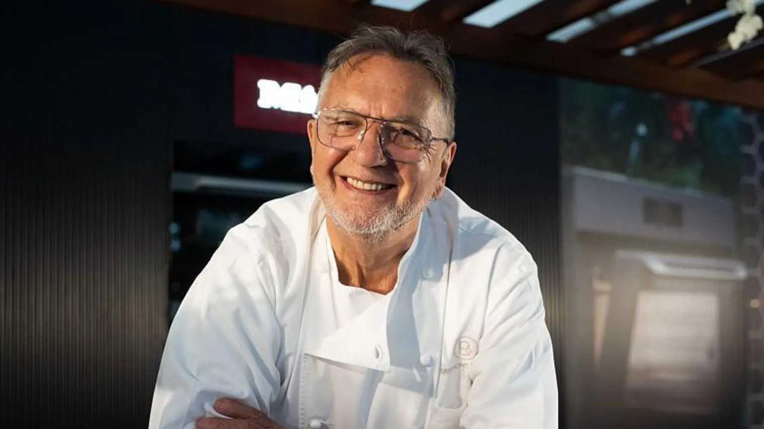  Raymond Blanc, wearing his chef's whites and glasses, smiles at the camera. He has short grey hair and a short beard, and his arms are folded across his chest