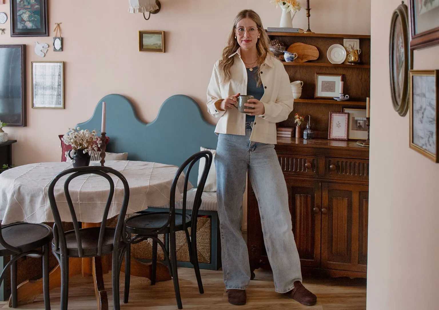 The Lovely Drawer Teri wearing blue jeans and a cream shirt unbuttoned over a blue vest top standing in a stylish dining room holding a mug