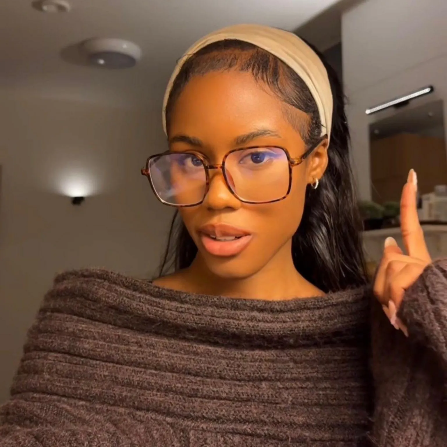 @ayoolaib A selfie of Ayoola wearing a brown off the shoulder knitted jumper and pointing to a wall light behind her