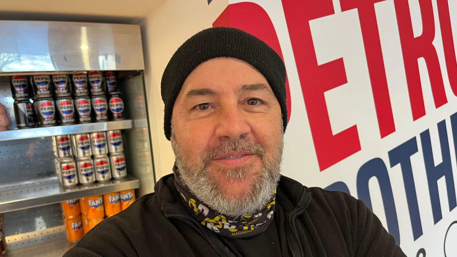 John Devine/BBC Mark Petrou has a grey beard and moustache and has a black woolly hat on, with a black, white and yellow snood around his neck, he has a black fleece top with black shirt under that, behind him are cold fizzy drinks in a chill cabinet.