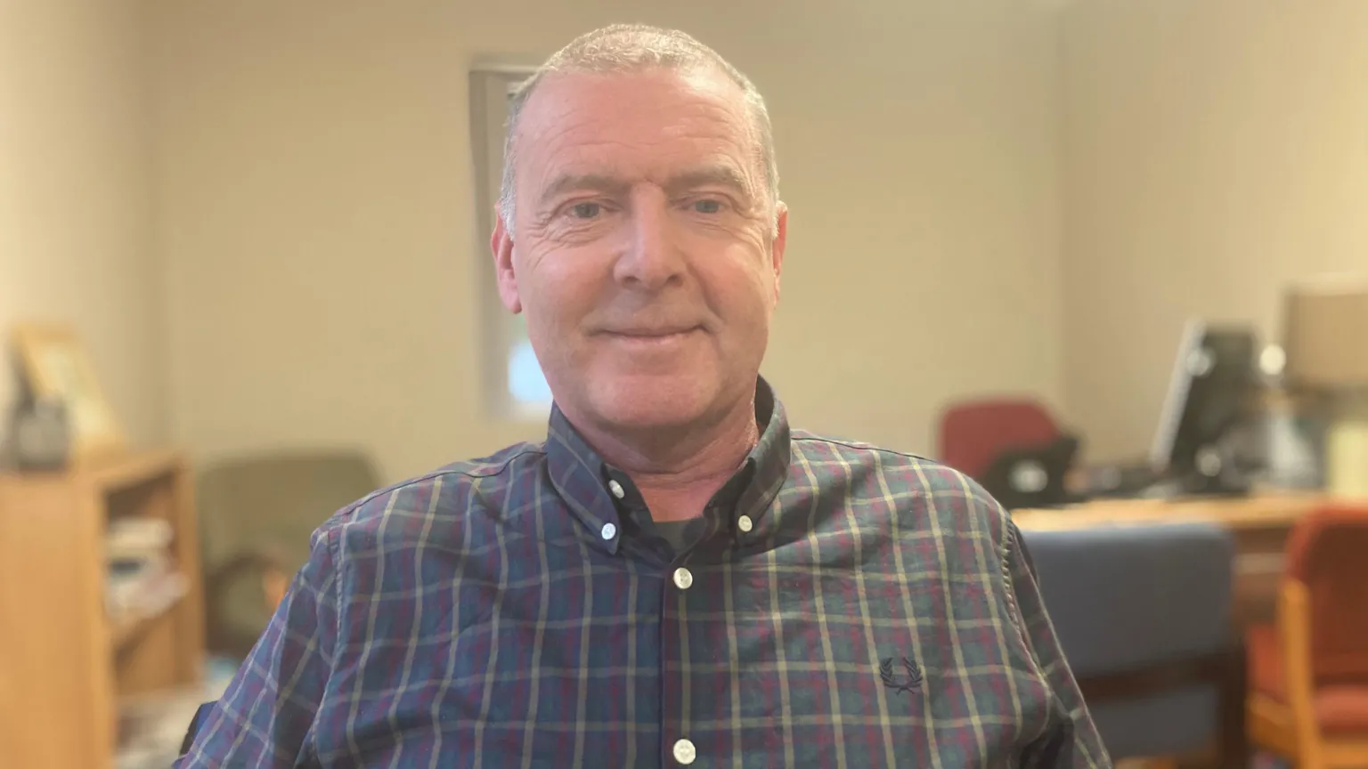 Dean is sitting in a room. He's wearing a checked shirt. The background is blurred. A table abd shelf are in the background.